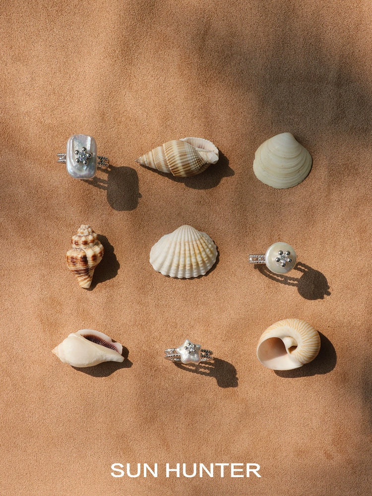 Elegant SUN HUNTER pearl riddle rings displayed among various seashells on a sandy background