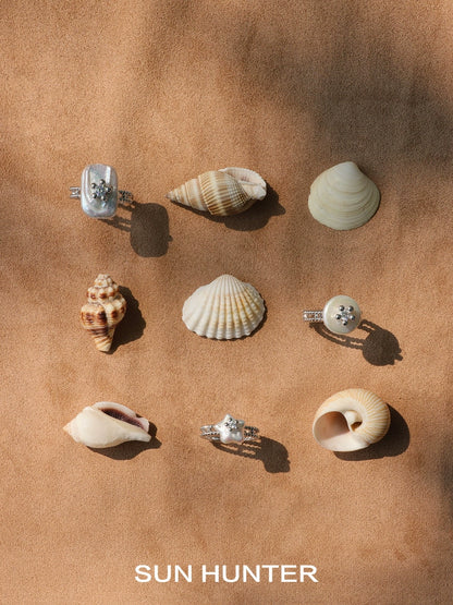 Elegant SUN HUNTER pearl riddle rings displayed among various seashells on a sandy background