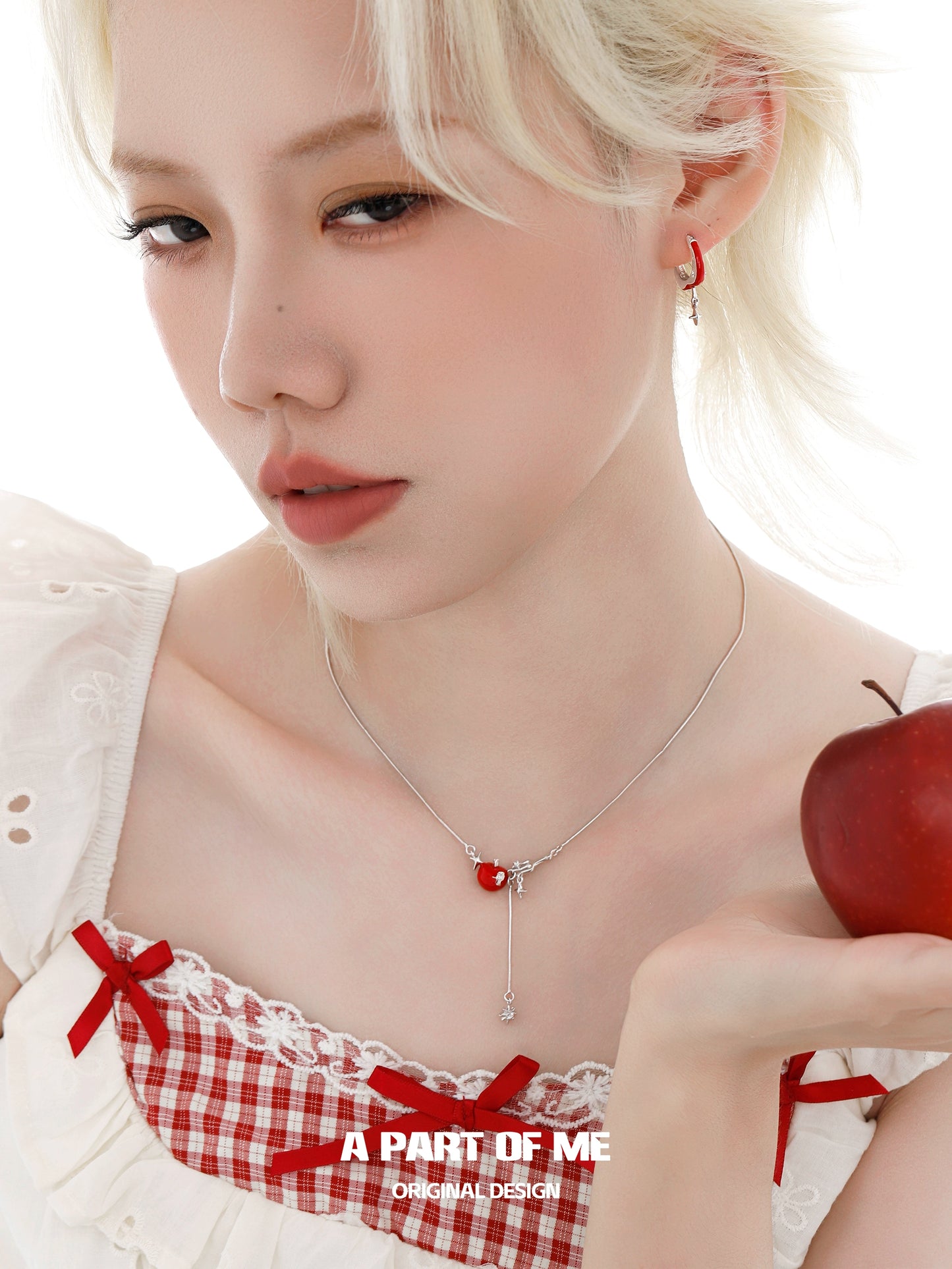 A woman wearing a red apple star necklace, holding an apple, in a stylish outfit with a delicate design