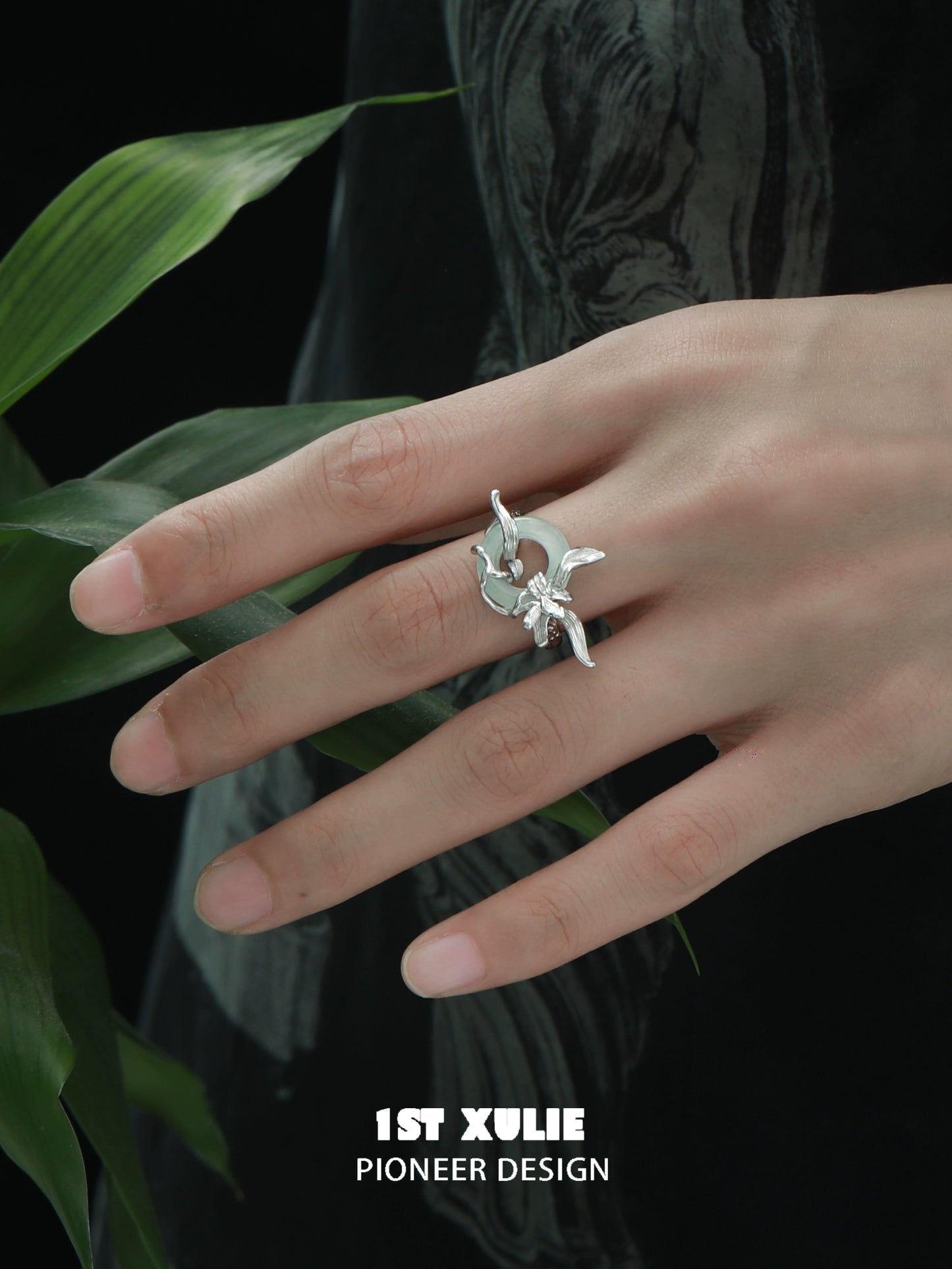 Silver iris safety buckle ring featured on a model's hand with leaves in the background