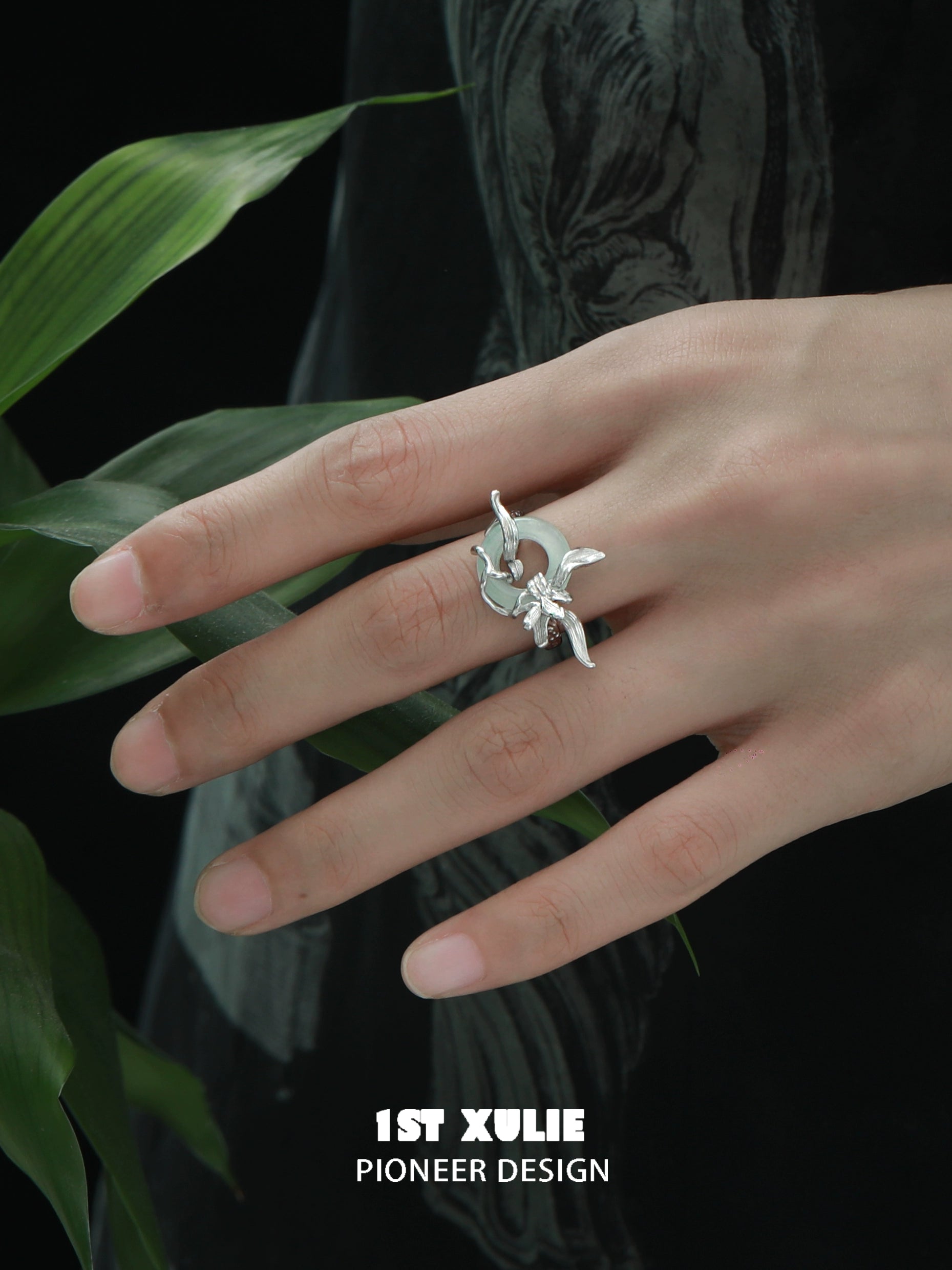 Silver iris safety buckle ring featured on a model's hand with leaves in the background