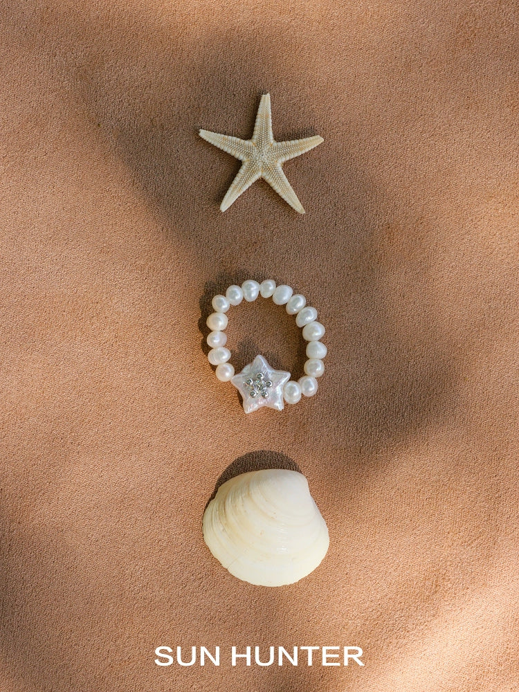 SUN HUNTER star pearl ring surrounded by a starfish and seashell on sandy surface